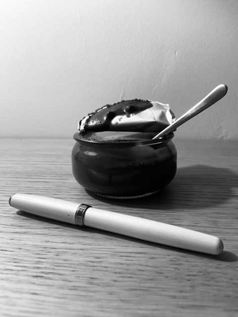 Photographie artistique en noir et blanc, vue de profil sur une table en bois clair. Au premier plan, un stylo blanc élégant est posé horizontalement. En arrière-plan, un petit pot de yaourt en verre foncé, dont l'opercule en aluminium est partiellement ouvert, contient une crème dessert nappée de chocolat. Une petite cuillère en métal est plantée à l'intérieur.